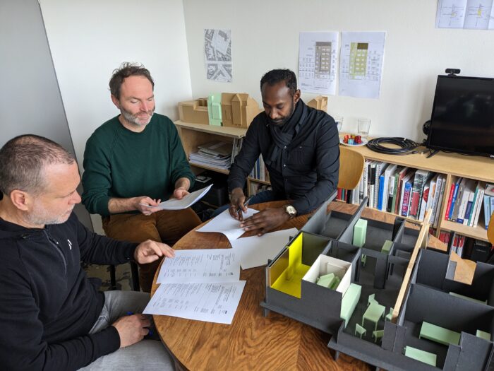 Drei Menschen sitzen um einen Tisch herum, auf dem ein Architektur-Modell von einem Stockwerk steht. Ein Mensch unterschreibt ein Dokument.