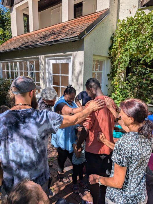 Menschen stehen beisammen und strecken ihre Hände in die Mitte. Die Hände berühren sich. Alle lächeln. Im Hintergrund ist ein Haus und eine Hecke zu sehen.