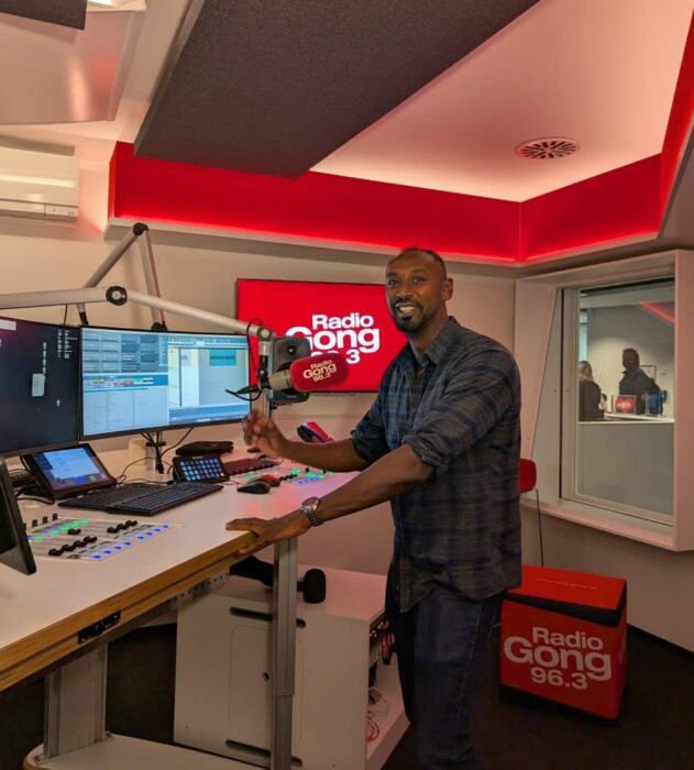 Taron ist zu sehen im Radio Studio des Radiosenders Gond 96.3. Er steht vor einem Bildschirm und einem Mikrofon. 